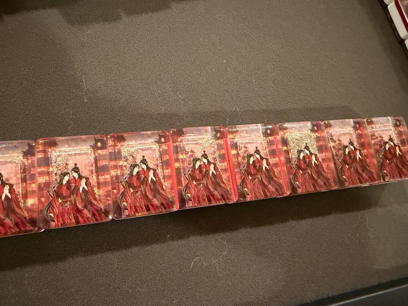 May include: A horizontal row of eight rectangular clear acrylic blocks, each featuring an identical printed illustration of two figures in traditional red robes standing together against a background of golden lanterns and architectural structures.