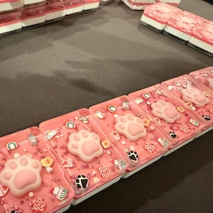 May include: Rectangular pink wax melts featuring a raised light pink cat paw print in the center. The surface is decorated with small colorful sprinkles, including tiny rainbows, flowers, and animal-themed shapes embedded in the wax.