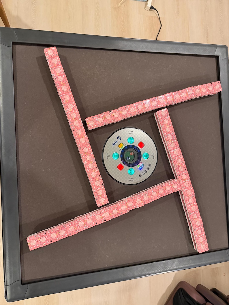 May include: A square automatic mahjong table with a dark brown felt surface and a black frame. Four rows of pink patterned mahjong tiles are arranged in a square formation around a central circular control panel with glowing green and red lights.