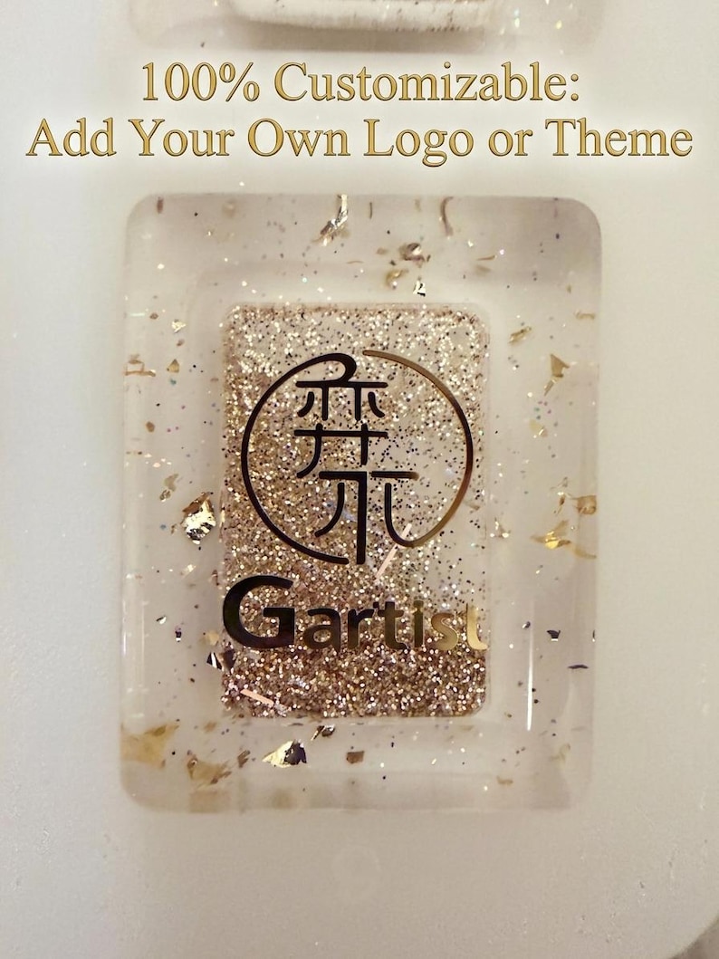 May include: A rectangular clear resin piece with gold glitter and metallic flakes embedded inside. The center features a circular logo with Asian characters and the text "Gartist" in black. Above the piece, the text reads "100% Customizable: Add Your Own Logo or Theme."