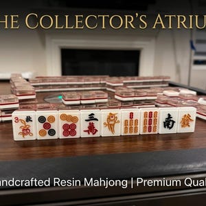 Può includere: Un set di tessere Mahjong in resina bianca con simboli e caratteri incisi colorati, disposte in file su una superficie in legno scuro. Le tessere presentano cime trasparenti e dettagli rossi, neri e dorati. Testo: THE COLLECTOR'S ATRIUM e Handcrafted Resin Mahjong | Premium Quality.