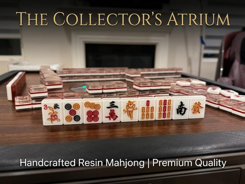 May include: A set of white resin Mahjong tiles with colorful engraved symbols and characters, arranged in rows on a dark wood-grain surface. The tiles feature clear tops and red, black, and gold details. Text reads THE COLLECTOR'S ATRIUM and Handcrafted Resin Mahjong | Premium Quality.