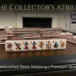 May include: A set of white resin Mahjong tiles with red, black, and gold engraved symbols and characters. The tiles are arranged in rows on a dark wood surface. The text THE COLLECTOR'S ATRIUM and Handcrafted Resin Mahjong | Premium Quality is visible.
