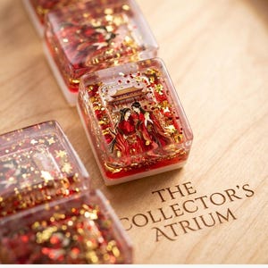 May include: A set of rectangular resin tiles featuring a clear top layer filled with red liquid, gold glitter, and a miniature illustration of two figures in traditional red robes standing before a building. The text THE COLLECTOR'S ATRIUM is visible.
