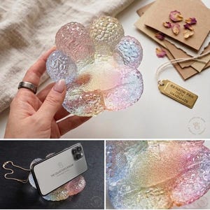 May include: A rainbow-colored, paw-shaped dish made of textured glass. The dish is held by a hand and has a tag that reads "RAINBOW PAW DISH - HANDMADE". The dish is shown holding a phone.