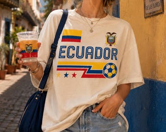 May include: An off-white short-sleeve t-shirt featuring the word ECUADOR in blue, a soccer ball graphic, the Ecuadorian flag, and three small stars. The shirt is paired with light blue denim shorts and a dark blue shoulder bag.