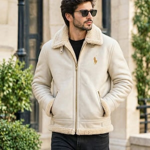 May include: A cream-coloured jacket with a tan shearling collar and lining, featuring a gold-tone zip closure, two side pockets, and an embroidered gold-tone logo on the left chest, worn over a black shirt and dark grey denim jeans.