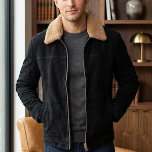 May include: A black suede jacket with a tan shearling collar and a gold zip. The jacket is worn with a grey sweater and dark wash denim jeans. The outfit is a classic, stylish look.