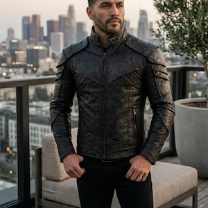 May include: A black leather jacket with a quilted pattern and shoulder accents. The jacket has a zip-up front and a stand-up collar. The person is wearing black pants. The background shows a city skyline.