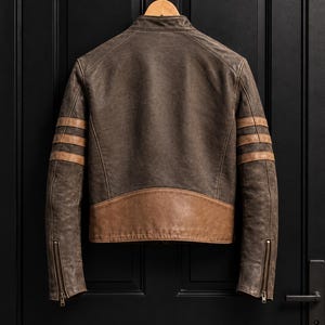 May include: A dark brown leather jacket featuring a stand-up collar, three horizontal tan stripes on each sleeve, and a tan leather panel across the lower back. The sleeves have metal zips at the cuffs, and the jacket is hung on a wooden hanger.