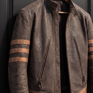 May include: A distressed brown leather jacket featuring a stand-up collar with a snap button closure, a full-length front zip, and two vertical zipped pockets. The sleeves are accented with three horizontal tan leather stripes.