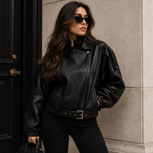 May include: A black leather jacket with a belted waist and a zip fastening. The jacket has a classic biker style with a high collar and a slightly oversized fit. The model is wearing black sunglasses and black trousers.