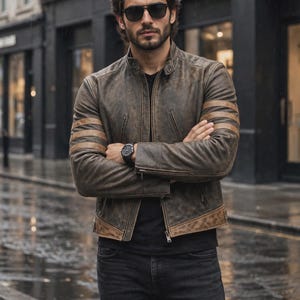 May include: A distressed brown leather jacket featuring a stand-up collar, a front zip fastening, and three horizontal stripes on each sleeve. The jacket is worn over a black t-shirt with black jeans, paired with dark sunglasses and a black wristwatch.