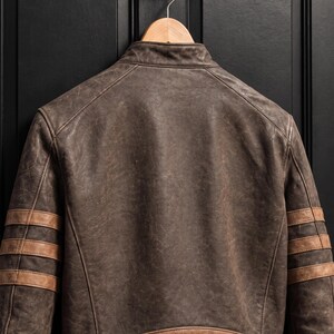 May include: A dark brown leather jacket featuring a distressed finish, a stand-up collar, and three horizontal tan stripes on each sleeve. The back of the jacket includes a solid tan leather panel along the bottom hem and visible stitching.