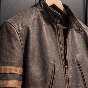 May include: A distressed dark brown leather jacket featuring a stand-up collar with a snap button closure, a central metal zip, and two vertical zipped pockets. The sleeve displays three horizontal light brown leather stripes.