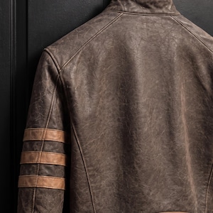 May include: A distressed dark brown leather jacket featuring a stand-up collar, three horizontal tan leather stripes on the upper sleeve, and a wide tan leather panel along the lower back hem, all constructed with visible reinforced stitching.
