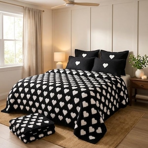 May include: A black bedspread and matching pillow shams featuring a repeating white heart pattern. The set includes a folded black and white heart-patterned throw blanket placed at the foot of the bed, creating a cohesive bedding ensemble.