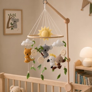 Può includere: Una giostrina per culla con struttura in legno e figure in feltro appese, tra cui un sole giallo, nuvole bianche, un canguro, un koala, un cacatua e un animale marrone, arricchita da foglie in feltro verdi e piccoli dischi di legno su fili bianchi.