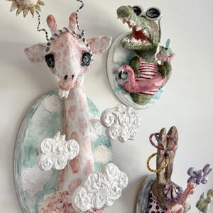 May include: Whimsical wall art featuring a giraffe, alligator, and rabbit. The giraffe is pink and white with cloud accents. The alligator wears sunglasses and a striped shirt. The rabbit has colorful accessories. Each piece is mounted on a painted oval base.