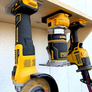May include: Three yellow and black DeWalt power tools hanging from a wooden shelf using custom mounts. The tools include an angle grinder with a grinding disc, a compact router labeled DCW600, and a reciprocating saw, all featuring 20V batteries.