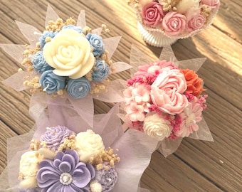 Puede incluir: Cuatro arreglos florales decorativos con flores de cera esculpidas en tonos rosa, blanco, azul y morado, acentuados con paniculata seca y envueltos en papel translúcido, presentados como ramos individuales sobre una superficie de madera.
