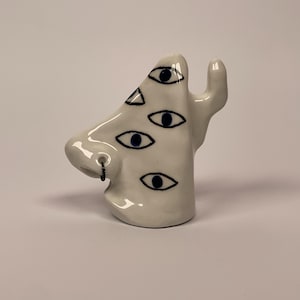 May include: A white ceramic sculpture shaped like a human nose with multiple hand-painted dark blue eyes scattered across the surface. The nose features a small, thin, dark metal wire ring inserted into the nostril area, adding a metallic detail.