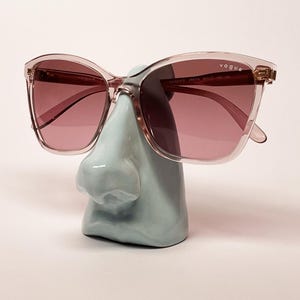 May include: Vogue sunglasses with translucent pink frames and tinted lenses. The brand name Vogue is printed in white on the upper right lens. The glasses are displayed on a light blue ceramic nose-shaped stand, showcasing the frame shape.