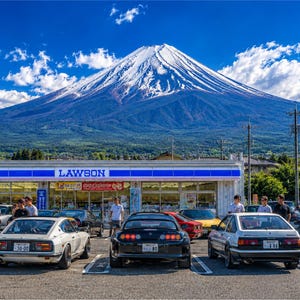 May include: A Lawson convenience store car park filled with various sports cars, including a black Toyota Supra, a white Datsun, and a white Toyota AE86, set against the backdrop of the snow-capped Mount Fuji under a bright blue sky.