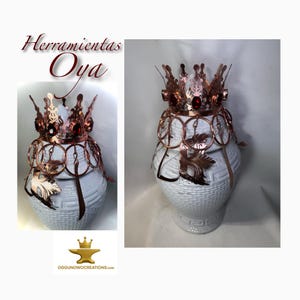 Peut inclure: Un vase en céramique blanc orné d'une couronne et d'éléments décoratifs en cuivre. La couronne est ornée de pierres précieuses rouges. L'image comprend le texte "Herramientas Oya" et le logo "OggunowoCreations.com".