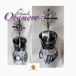 May include: A decorative metal crown with the text "Obatala Obamoro". The crown is silver with a sunburst design on top and a series of ladders and figures around the base. The crown is displayed on a white form.