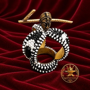 May include: A circular napkin ring featuring a gold-toned metal base adorned with alternating black and white beads. A gold-toned metal ornament shaped like a stylized face is attached to the top, positioned horizontally across a thin bar.