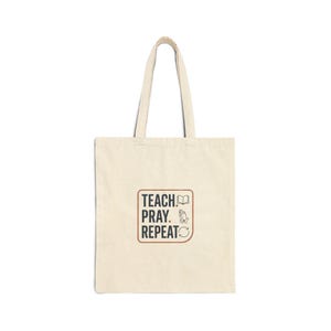 May include: A cream-coloured tote bag featuring the text "TEACH, PRAY, REPEAT" in a brown font within a rectangular design. The design includes a book, praying hands, and a recycling symbol. The bag has two handles.