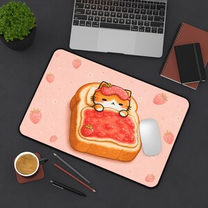May include: A rectangular desk mat featuring a light pink background with a repeating pattern of small strawberries. The central graphic depicts a slice of toast topped with red jam and a small orange cat wearing a strawberry-patterned sleep mask.