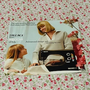 May include: A vintage advertisement for the Silvia sewing machine from Necchi of Italy. The print features a person in a beige sweater operating a black sewing machine, with a child nearby. Text reads: The new sewing machine that does more than just sew.