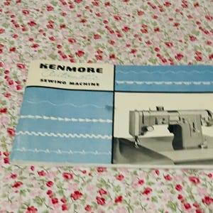 Könnte beinhalten: Ein rechteckiges Handbuch für die Kenmore Automatic Nähmaschine mit einem weiß-blau-schwarzen Einband. Der Einband zeigt den Text Kenmore Automatic Sewing Machine, Abbildungen verschiedener Stichmuster und eine schwarz-weiße Nähmaschine.