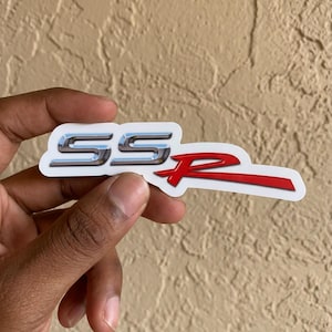 May include: A chrome and red SSR emblem sticker. The sticker features the letters "SS" in chrome and the letter "R" in red. The sticker is held in a hand against a beige background.