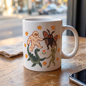 May include: A white ceramic mug featuring a colorful illustration of a beige Bigfoot, a black Mothman, a green Loch Ness monster, and a green alien holding hands in a circle. The design includes small orange stars and a single orange flower.