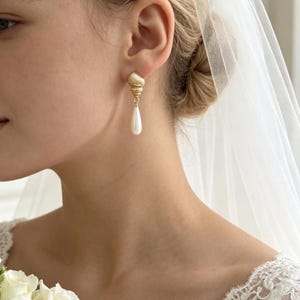 Puede incluir: Un pendiente dorado con un poste en forma de concha espiral texturizada, del cual cuelga un colgante de perla blanca lisa en forma de lágrima. El pendiente se lleva en el lóbulo de la oreja, con un velo blanco transparente visible al fondo.