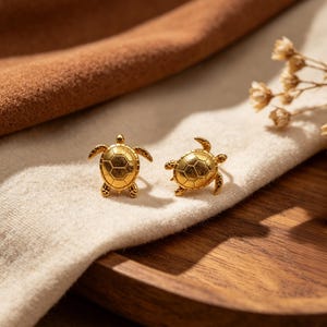 Peut inclure: Une paire de boucles d'oreilles à clous dorées en forme de tortues de mer. Chaque boucle présente une carapace texturée et détaillée avec une finition polie, reproduisant la forme distincte d'une tortue avec quatre nageoires et une petite tête.