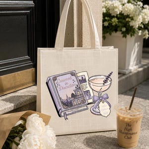 May include: A cream-colored canvas tote bag featuring a graphic print of an open book titled "Crowned in Discretion" next to a stemmed cocktail glass with a lavender sprig and a purple ribbon tag. The bag has two top handles and a grid pattern.
