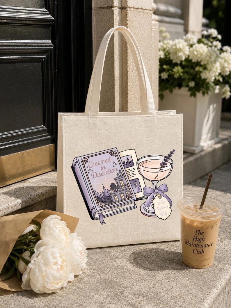 May include: A cream-colored canvas tote bag featuring a graphic print of an open book titled "Crowned in Discretion" next to a stemmed cocktail glass with a lavender sprig and a purple ribbon tag. The bag has two top handles and a grid pattern.