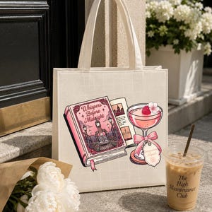 May include: A cream-colored canvas tote bag featuring a graphic print of an open book titled Whispers Before Midnight and a pink cocktail glass with a raspberry garnish. The bag has two top handles and a subtle grid pattern on the fabric surface.