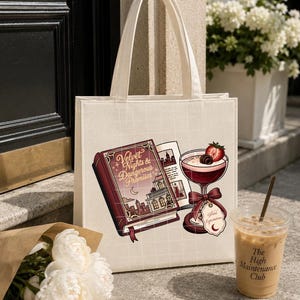 May include: A cream-colored canvas tote bag featuring a graphic print of a red book titled Velvet Nights & Dangerous Promises, an open book, and a cocktail glass garnished with a strawberry and chocolate, all set against a faint white grid pattern.