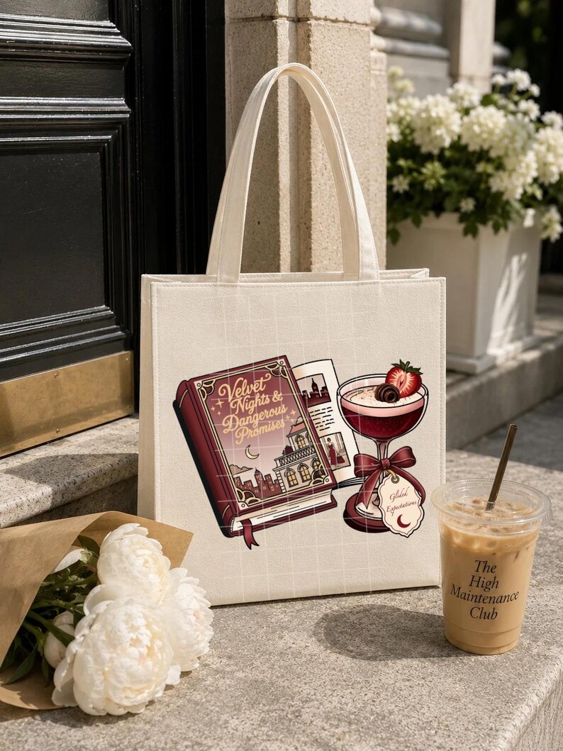 May include: A cream-colored canvas tote bag featuring a graphic print of a red book titled Velvet Nights & Dangerous Promises, an open book, and a cocktail glass garnished with a strawberry and chocolate, all set against a faint white grid pattern.
