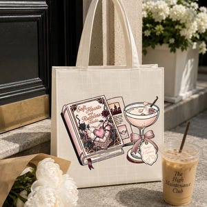 May include: A cream-colored canvas tote bag featuring a graphic print of an open book titled Soft Hearts and Ruthless Taste, a cocktail glass with a pink bow, and a tag labeled Savage Elegance, all rendered in shades of pink, black, and white.