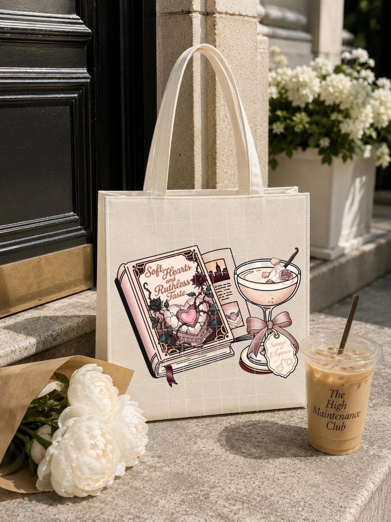 May include: A cream-colored canvas tote bag featuring a graphic print of an open book titled Soft Hearts and Ruthless Taste, a cocktail glass with a pink bow, and a tag labeled Savage Elegance, all rendered in shades of pink, black, and white.