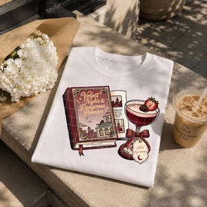 May include: A white t-shirt featuring a graphic print of a maroon book titled Velvet Nights & Dangerous Promises, an open book page, and a cocktail glass garnished with a strawberry and a chocolate, with a tag labeled Great Expectations.