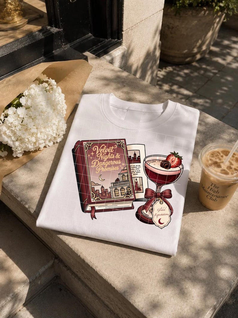 May include: A white t-shirt featuring a graphic print of a maroon book titled Velvet Nights & Dangerous Promises, an open book page, and a cocktail glass garnished with a strawberry and a chocolate, with a tag labeled Great Expectations.
