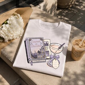 May include: A white t-shirt featuring a graphic illustration of a purple book titled Crowned in Discretion, a cocktail glass with a lavender sprig, a purple ribbon bow, and a tag labeled Velvet Defiance with a small crown icon printed on the front.
