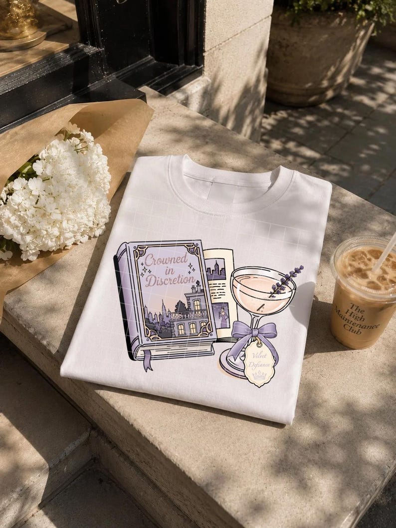 May include: A white t-shirt featuring a graphic illustration of a purple book titled Crowned in Discretion, a cocktail glass with a lavender sprig, a purple ribbon bow, and a tag labeled Velvet Defiance with a small crown icon printed on the front.